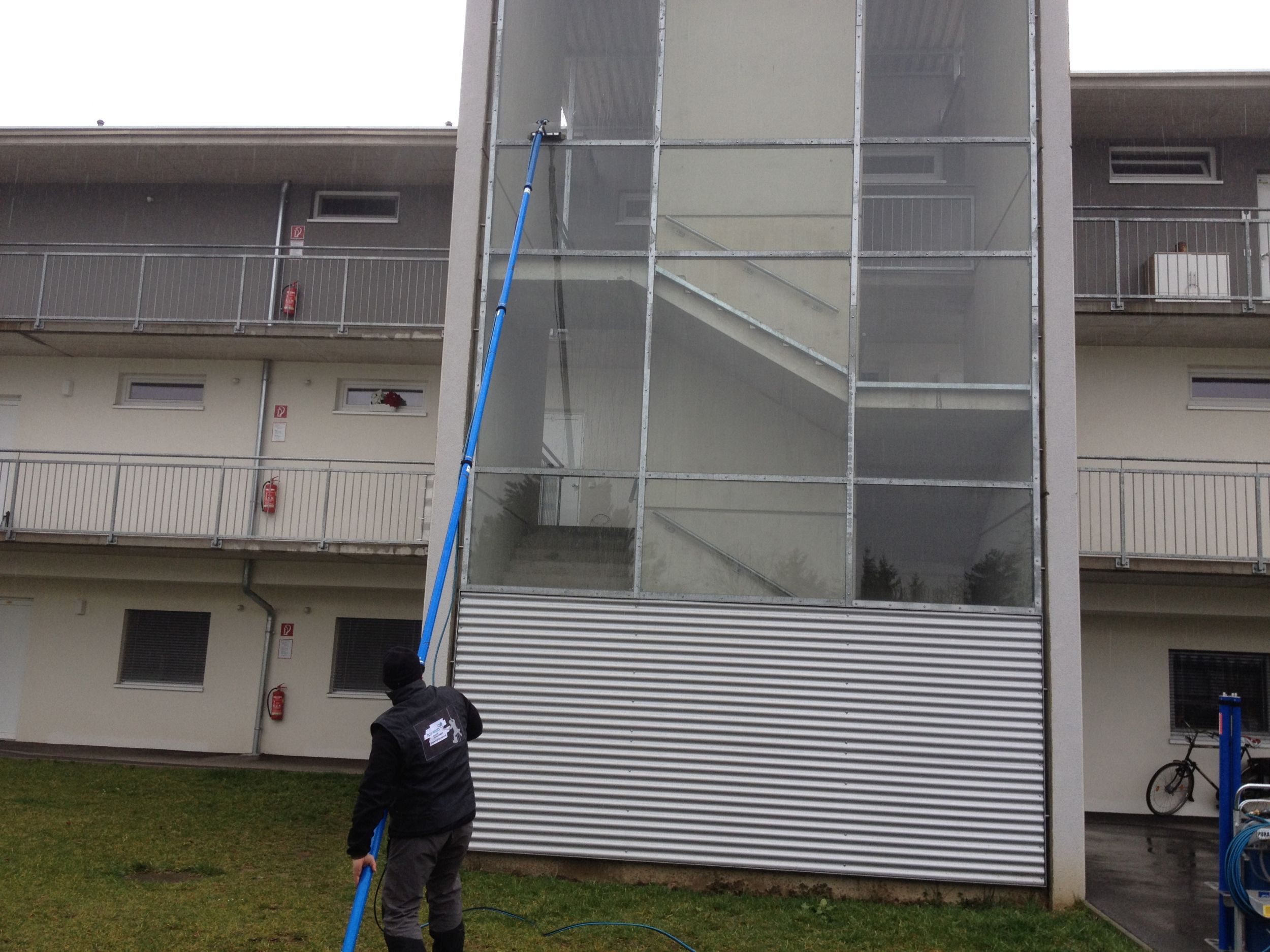 Ein H2Concepts Mitarbeiter reinigt die Fenster eines mehrstöckigen Gebäudes mit einem langen Teleskopstiel.