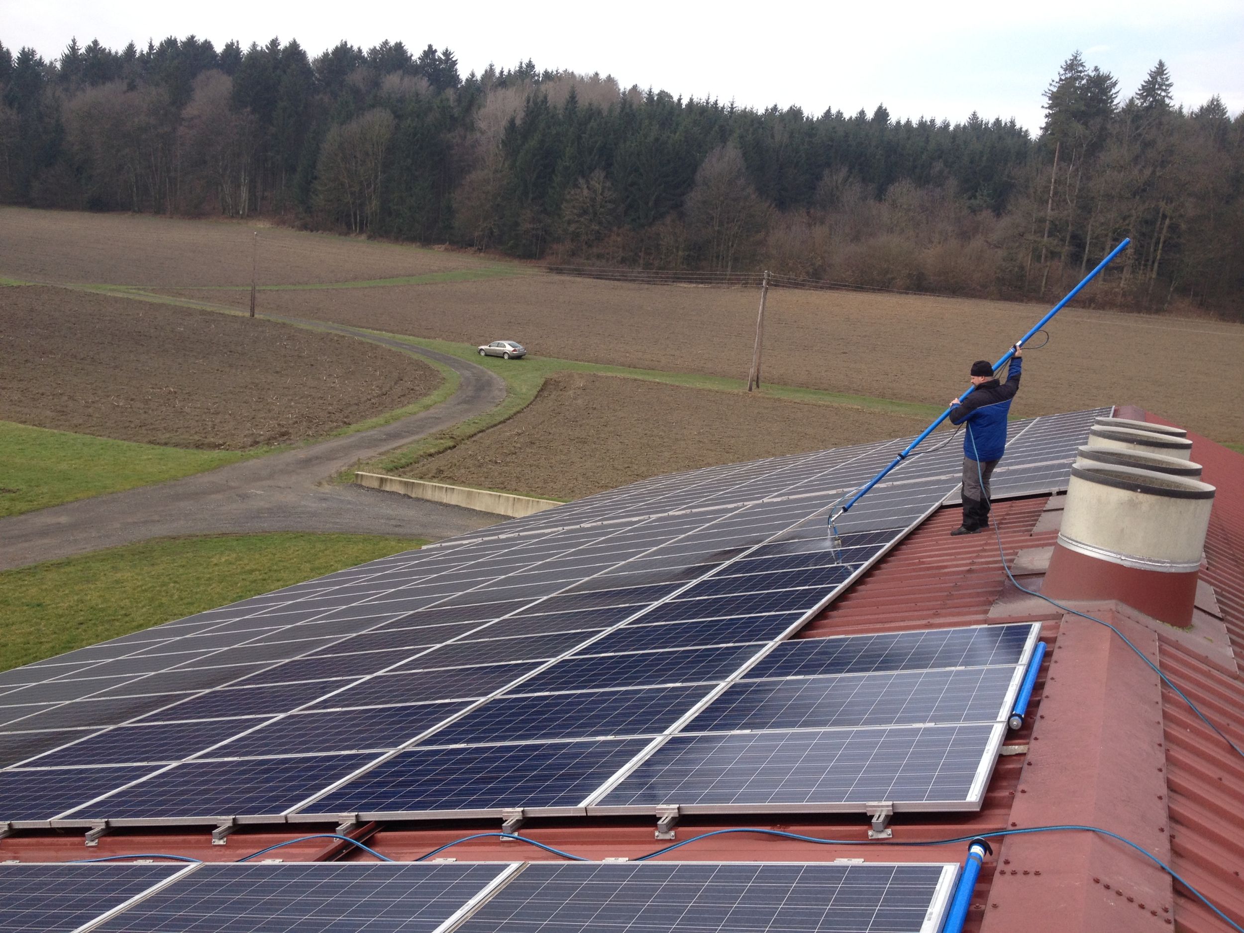 Person reinigt Solarpanels auf einem Dach mit einem langen Besenstiel.