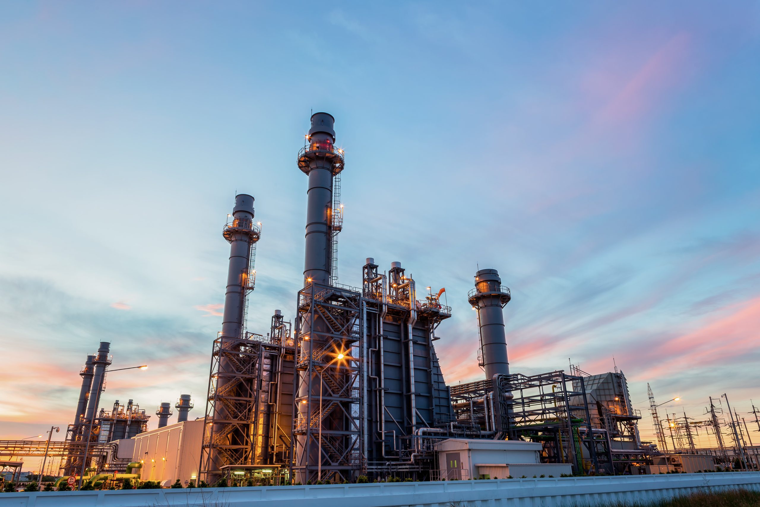 Große Industrieanlage mit mehreren hohen Schornsteinen und Rohrleitungen, beleuchtet bei Sonnenuntergang. Der Himmel zeigt sanfte Blau- und Rosatöne.