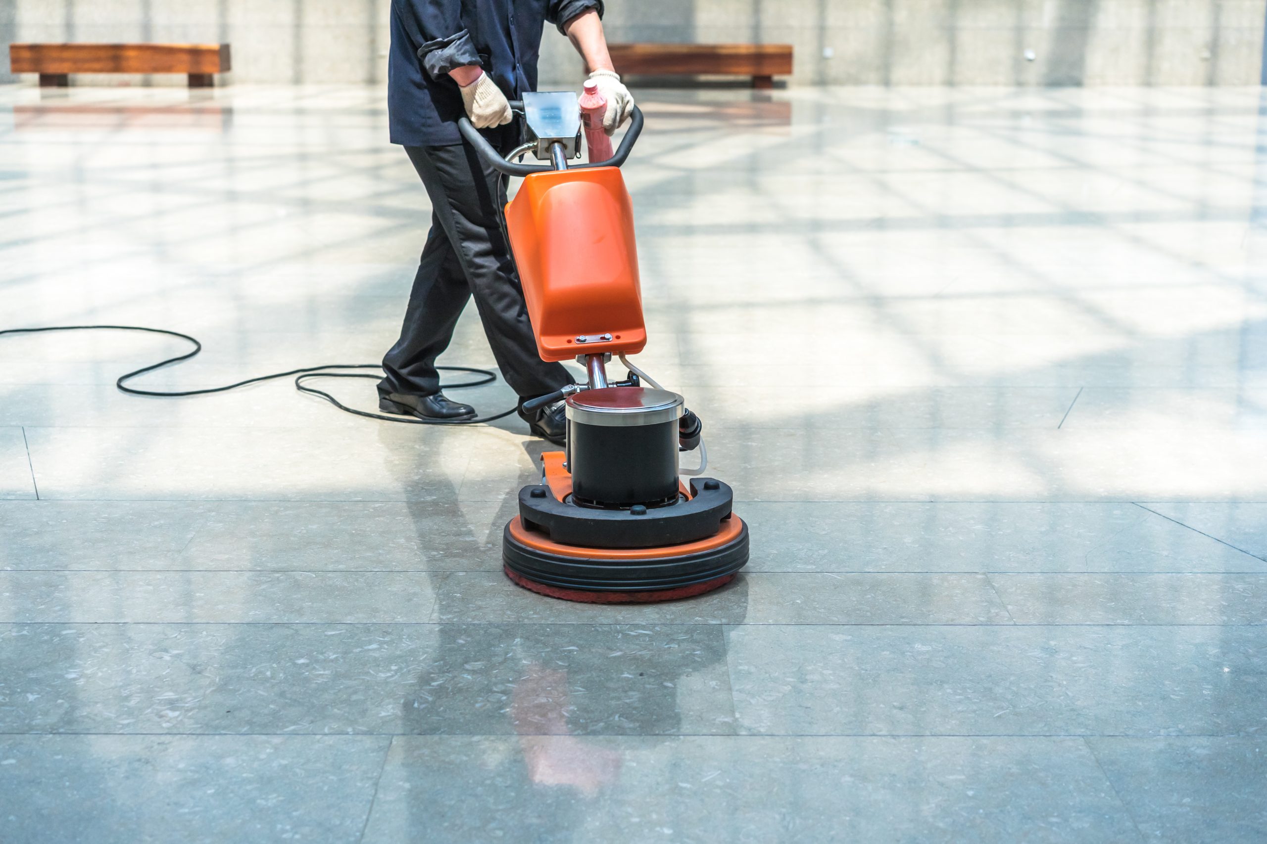 Reinigungskraft poliert einen glänzenden Steinboden mit einer orangefarbenen Poliermaschine in einem großen, lichtdurchfluteten Raum.