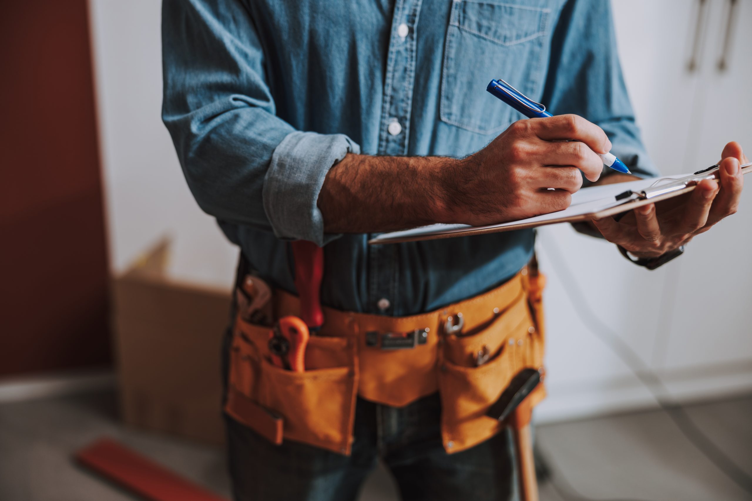 Handwerker in blauer Jeansjacke, der Notizen auf einem Block macht, mit Werkzeuggürtel um die Taille.
