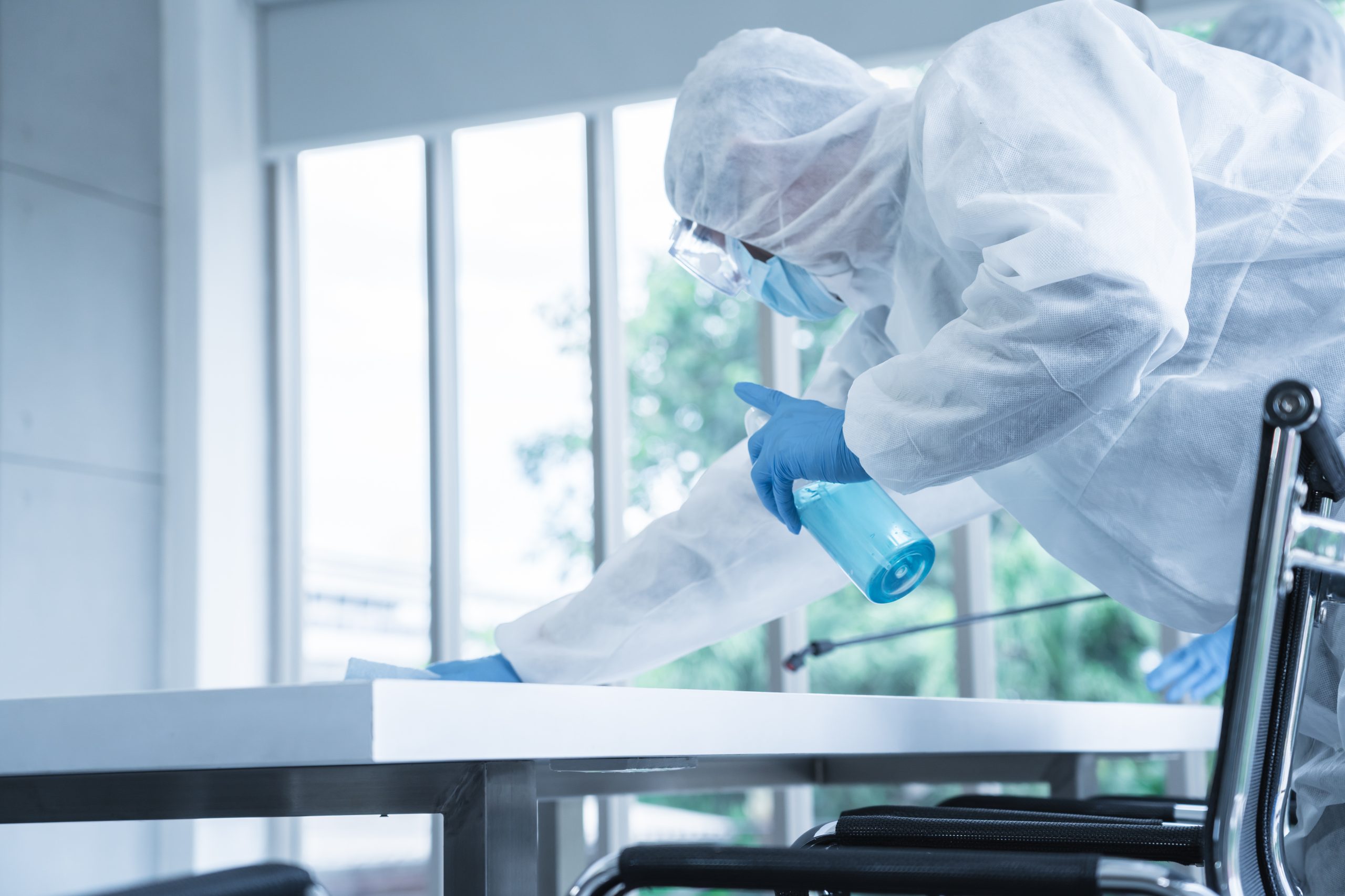 Person in Schutzanzug, Handschuhen und Maske desinfiziert einen Tisch mit Reinigungsmittel in einer hellen, modernen Umgebung.