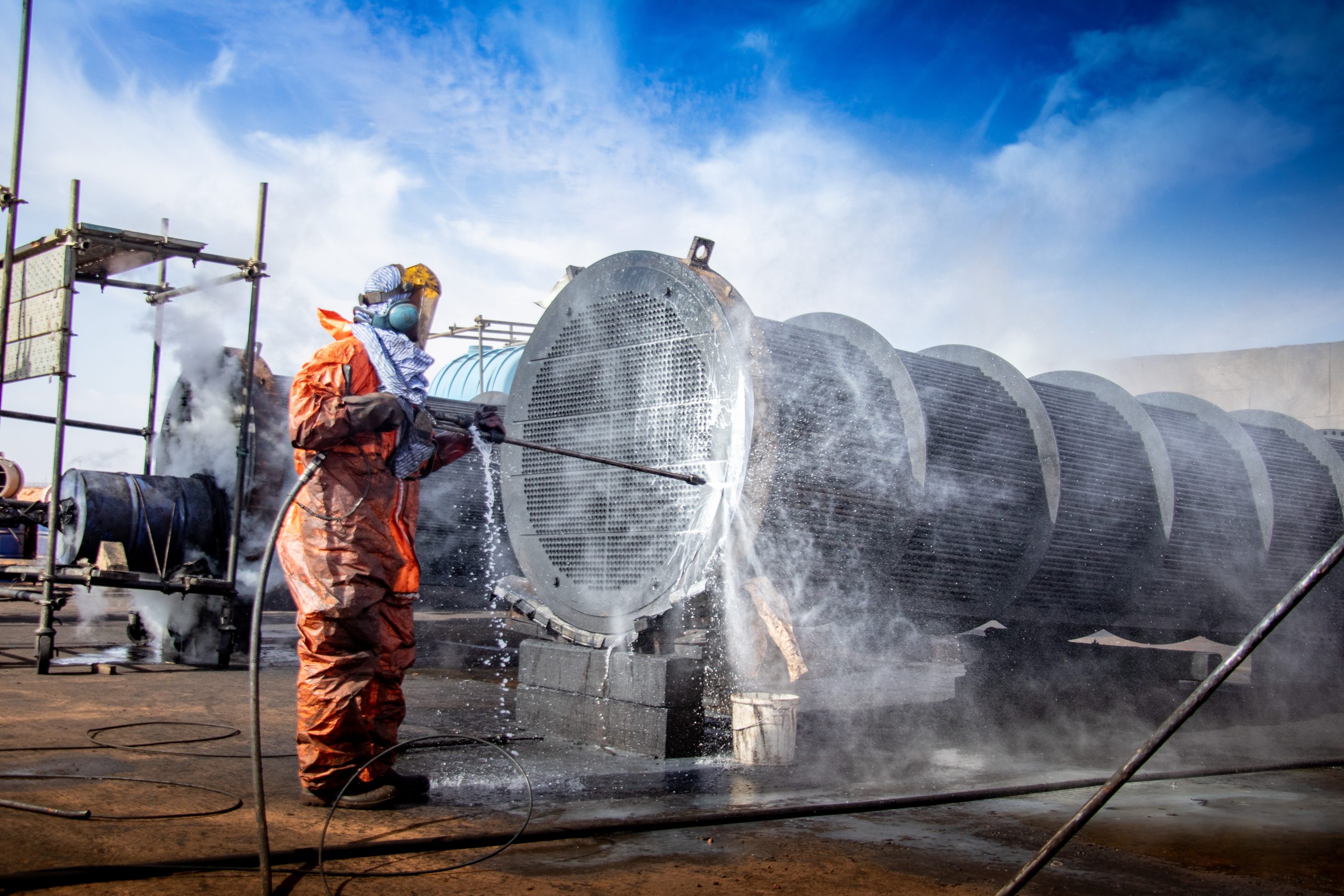 Arbeiter in Schutzkleidung reinigt ein großes Metallbauteil mit einem Hochdruckreiniger. Wasser und Schaum spritzen, während Dampf aufsteigt.
