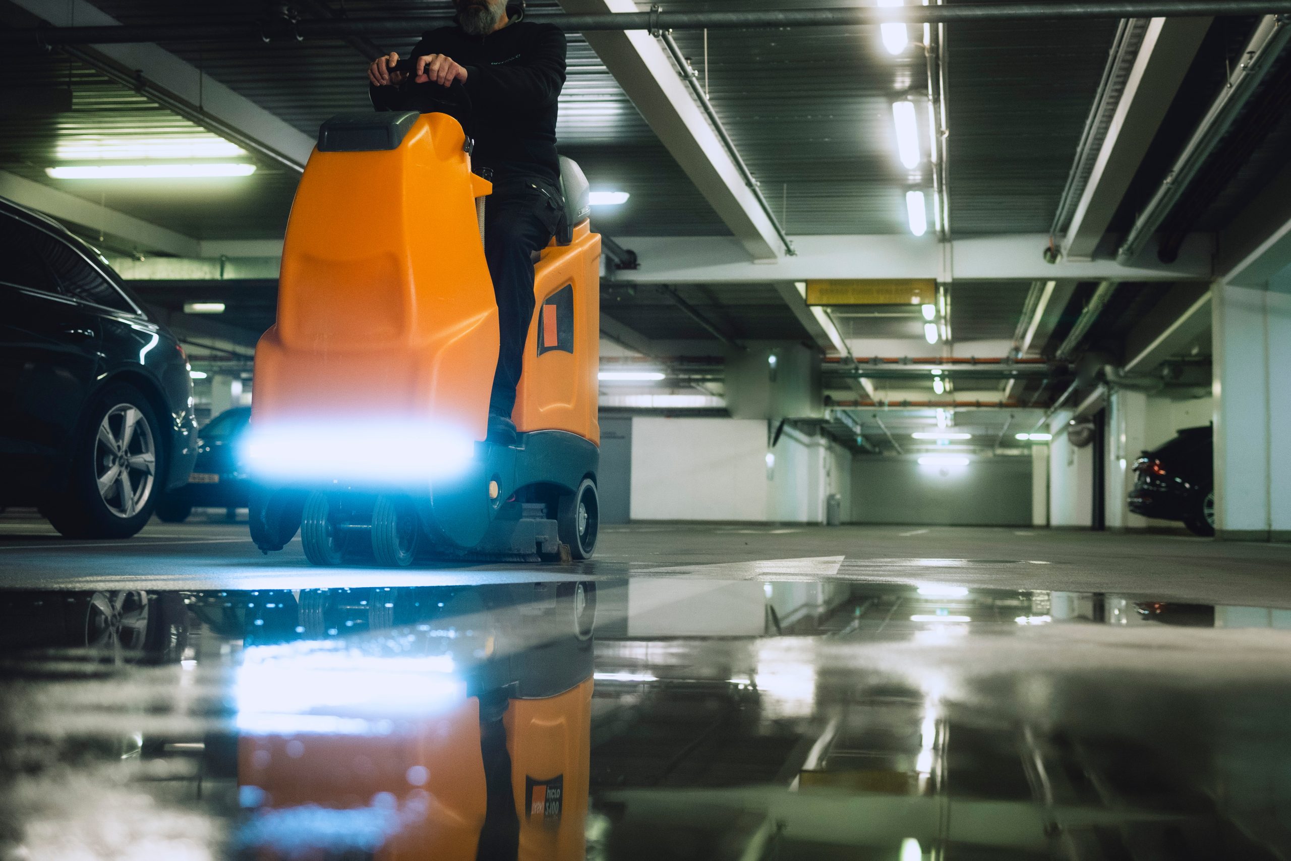 Eine orange Bodenreinigungsmaschine wird in einer Tiefgarage verwendet, mit reflektierendem Wasser auf dem Boden.